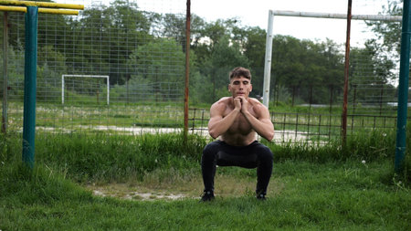 Male shirtless athlete doing squats at sports ground. Young sportsman working at summer nature....