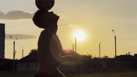 Young man juggling soccer ball on stadium at sunset. Professional footballer kicking ball at green field. Sportsman practicing tricks at meadow with sunlight at background. Freestyle footballの写真素材
