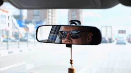 Eyes in sunglasses of male driver reflecting in rearview mirror while riding motor auto. Young guy...