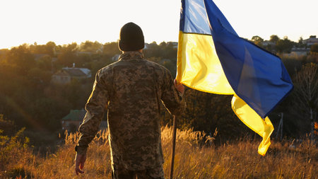 Soldier of ukrainian army going with lifting blue-yellow banner at sunset. Young male military in uniform walks with waving flag of Ukraine at countryside. Victory against russian aggression conceptの写真素材