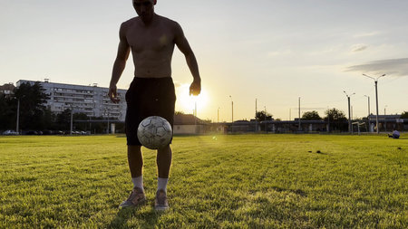 Young man juggling soccer ball on stadium at sunset. Professional footballer kicking ball at green field. Sportsman practicing tricks at meadow with sunlight at background. Freestyle footballの写真素材