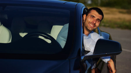 Young smiling entrepreneur in shirt leaning out of driver window at modern electric car and looking...