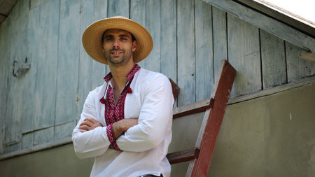 Portrait of smiling guy in straw hat against the background of his house. Young ukrainian man wearing an embroidered shirt looking into camera outdoor. Concept of national identity and patriotismの写真素材