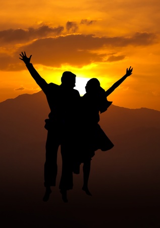 Silhouette of loving couple jumping on a hill against the sun and mountains rangeの写真素材