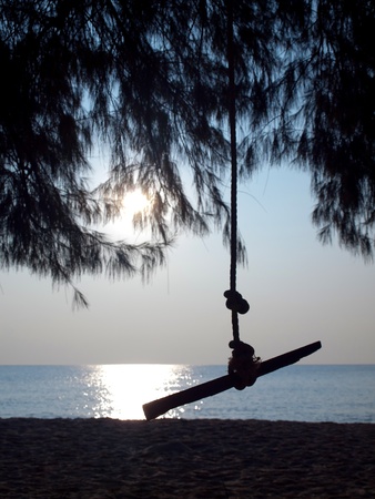 Silhouette swing in shady of pine against beach sea and sunの写真素材