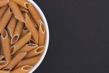 White glass bowl of raw penne rigate pasta. Isolated on a dark gray background. Top view close upの写真素材
