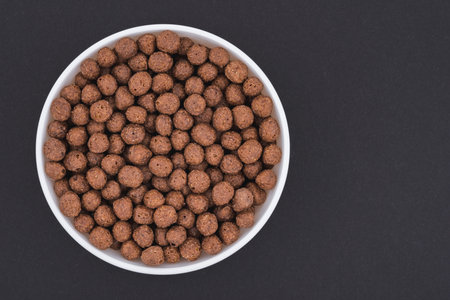 White glass bowl of chocolate corn balls. Isolated on a dark gray background. Top view close up