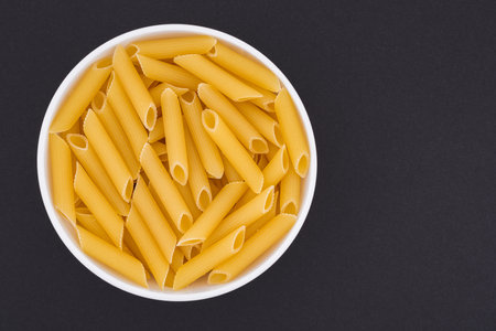 White glass bowl of raw penne rigate pasta. Isolated on a dark gray background. Top view close upの写真素材