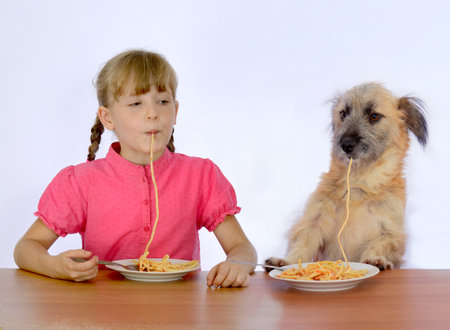 little girl with dog eating pasta sitting at the table on white backgroundの写真素材