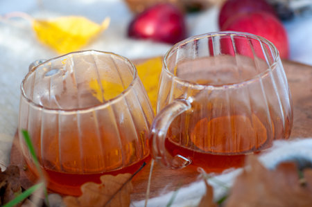 Two cups of tea sit on a wooden table with leaves and apples. The cups are clear and the tea is brownの写真素材
