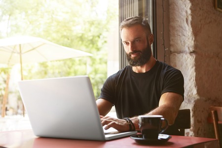 Handsome Bearded Businessman Wearing Black Tshirt Working Laptop Wood Table Urban Cafe.Young Manager Work Notebook Modern Interior Design Place.Coworking Process Business Startup.Blurred Backgroundの写真素材