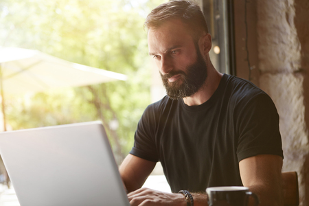 Concentrated Bearded Man Wearing Black Tshirt Working Laptop Wood Table Urban Cafe.Young Manager Work Notebook Modern Interior Design Loft Place.Coworking Process Business Startup.Blurred Backgroundの写真素材