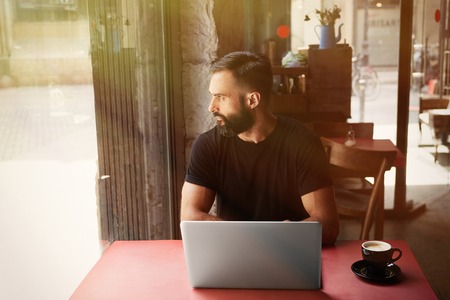 Young Bearded Businessman Wearing Black Tshirt Working Laptop Urban Cafe.Man Sitting Wood Table Cup Coffee Looking Through Window.Coworking Process Business Startup.Blurred Background.Sunlight effectの写真素材