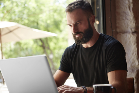 Concentrated Bearded Man Wearing Black Tshirt Working Laptop Wood Table Urban Cafe.Young Manager Work Notebook Modern Interior Design Loft Place.Coworking Process Business Startup.Color Filterの写真素材