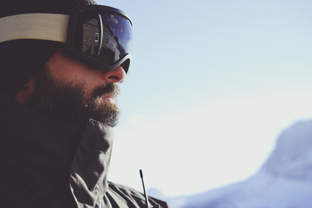 Close-up portrait of bearded young snowboarded in sunglass mask, at the ski resort on the background of mountains and blue sky.Blurred background.Horizontal.の写真素材