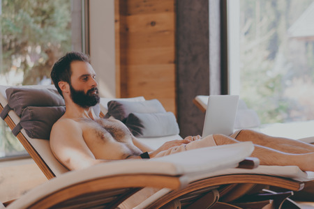 Handsome bearded Man using laptop computer while resting in spa hotel. Man relaxing after hard day at spa salon.Sunligth wooden interior. Cropped.の写真素材