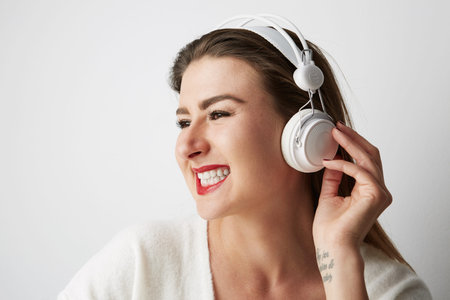 Smiling beautiful young woman listening to music on headphones over empty light background at...