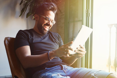 Young happy bearded man sitting home and using digital tablet for surfing web internet. Man using gadget at apartment. Horizontal. Blurred backgroundの写真素材