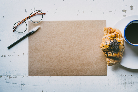 Mockup blank craft sheet of blank craft paper, pen, eye glasses and morning coffee cup with croissant on white wooden desk. Business empty background for letter writing.Creative work conceptの写真素材