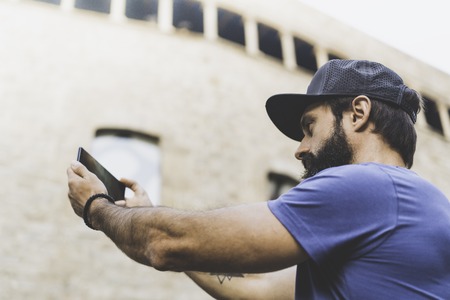 Bearded muscular man wearing black snapback cap and making selfie with his own smartphone. Man walking on street and using mobile.Blurred background. Horizontalの写真素材