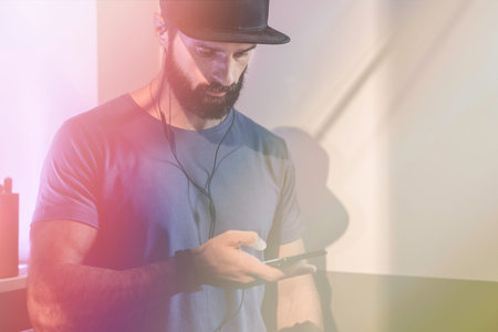 Bearded pensive man wearing black snapback cap standing on cafe with headphones. Men using mobile...