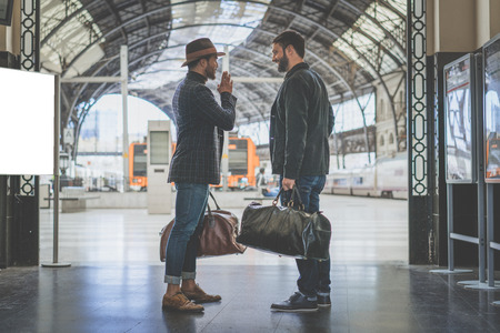 Two confident multi-ethnic bearded businessmans together wearing casual clothes and holding travel bags in hands waiting the train on the railway platform.の写真素材