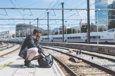 Attractive confident hispanic man having a backpack front of him with a clothes bag, walking beside a platform at train stationの写真素材