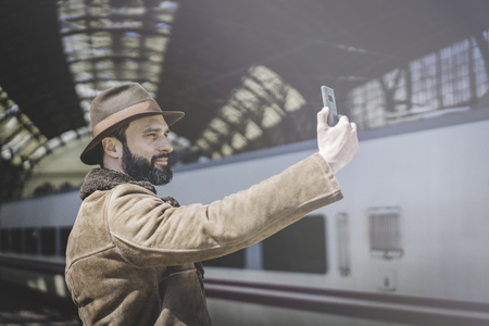 Attractive adult businessman making selfie on a mobile phone.Casual professional entrepreneur using smartphone outside while waiting train on railway stationの写真素材