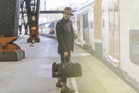 Handsome confident hispanic hipster with travel bag wearing casual clothes and hat waiting the train on the railway platform.の写真素材
