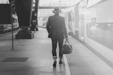 Young confident hispanic business man with travel bag wearing casual clothes and hat looking the train on the railway platform. Black and whiteの写真素材
