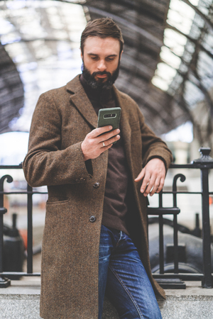 Confident bearded businessman standing with travel bag at the hall of railway station and texting message on his mobile phoneの写真素材