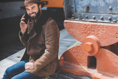 Attractive bearded adult caucasian man holding smart phone in his hand and calling while he standing at the railway platform waiting for the train.の写真素材