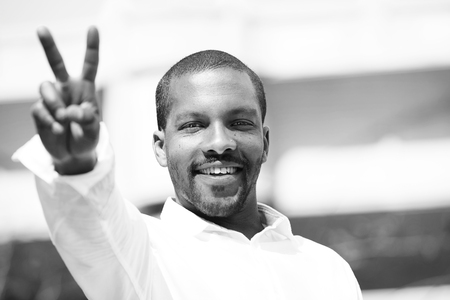 Portrait of confident young African-American man hipster in white shirt showing fingers symbol V.Blue sky with clouds on background. Black and white.の写真素材