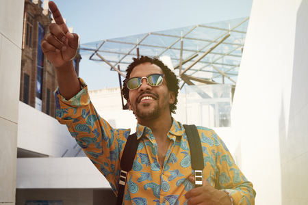 Portrait of attractive young African-American hipster in casual wear and sunglasses smiling and pointing hand.Blurred backgroundの写真素材