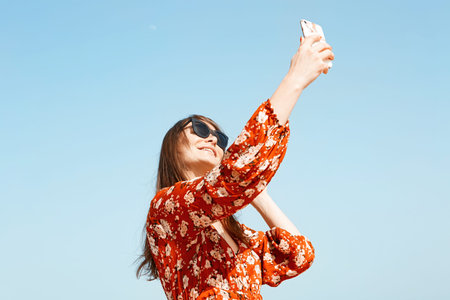 Happy young girl hipster taking selfie outdoors of city. Blue clear sky on blurred background