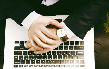Working outside office.Young businessman in formalwear typing on laptop and checking hand watch while sitting at city green grass outdoor. Closeup top viewの写真素材