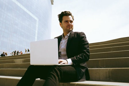 Attractive businessman wearing suit and using modern laptop outdoors. Successful banker working outside during breakの写真素材