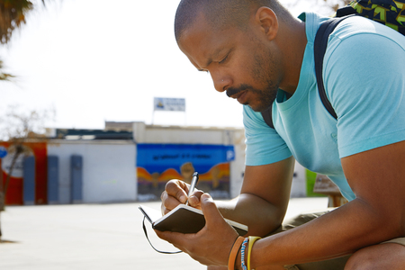 Attractive pensive African-American hipster man wearing backpack and making notes in copybook in city park. Student preparing for lesson at collegeの写真素材