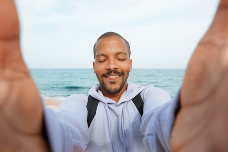 Handsome young African-American hipster man making smile and taking hand selfie with closed eyes outdoors of cityの写真素材