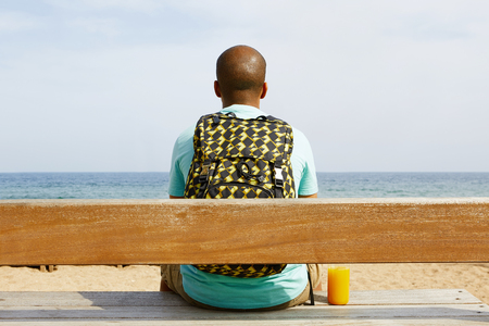 Attractive young African-American hipster wearing t-shirt and sitting on bench at summer day on the beach with backpackの写真素材
