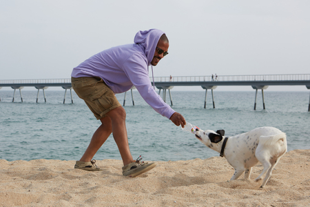 Smiling young African-American man hipster in sport hoody playing with his dog on the beach at sunny dayの写真素材