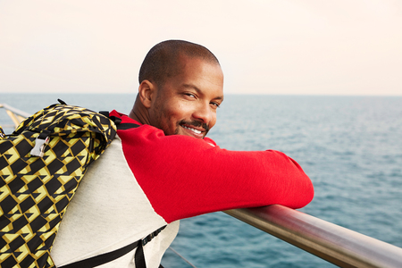 Happy young African-American hipster man wearing backpack dreaming on the beach and enjoying sea view.Closeup portrait.の写真素材