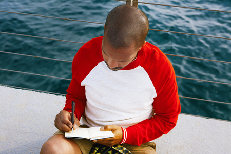 handsome young African-American hipster man relaxing on the beach and making text notes in paper notebook. Blue sea on the blurred background.Croppedの写真素材