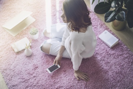Happy young beautiful woman using electronic gadgets while sitting on the floor in the living roomの写真素材