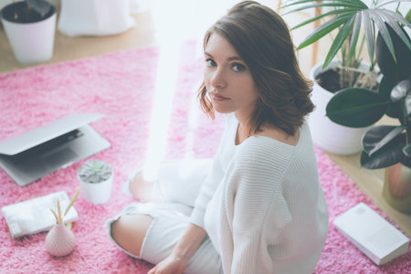 Beautiful woman sitting on pink carpet on the floor at modern and light coloured living room with...