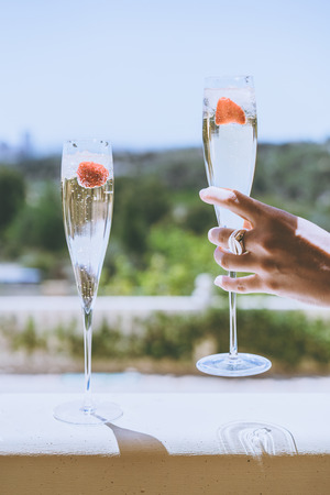 Female hand holding a glasses champagne with strawberry inside on sunny terrace overlooking beautiful view at summer day outside of the cityの写真素材