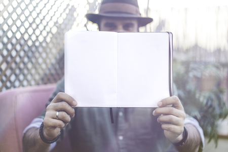 Attractive Hipster blogger holding notepad with open blank sheets on hands, while spending time on terrace of cafe.Space for Copy paste text. Sunlight effectの写真素材