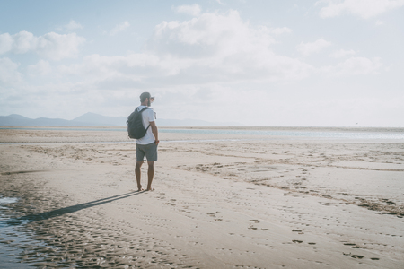 Attractive muscular bearded man standing on the sea shore at sunrise with backpack and looking horizonの写真素材