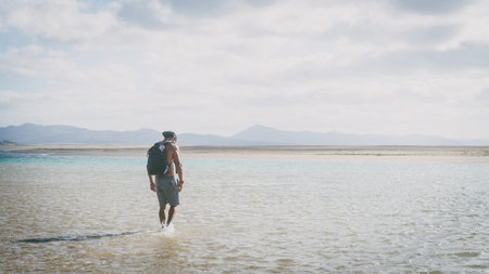 Young muscular bearded man walking on the sea shore at sunrise with backpack. Wide