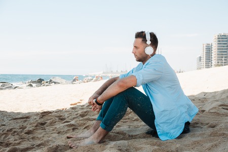 Young hipster man in a blue shirt and jeans listening to music in headphones on a smartphone and is sitting on beach sand looking at sea. Digital Music lounge and relaxing conceptの写真素材